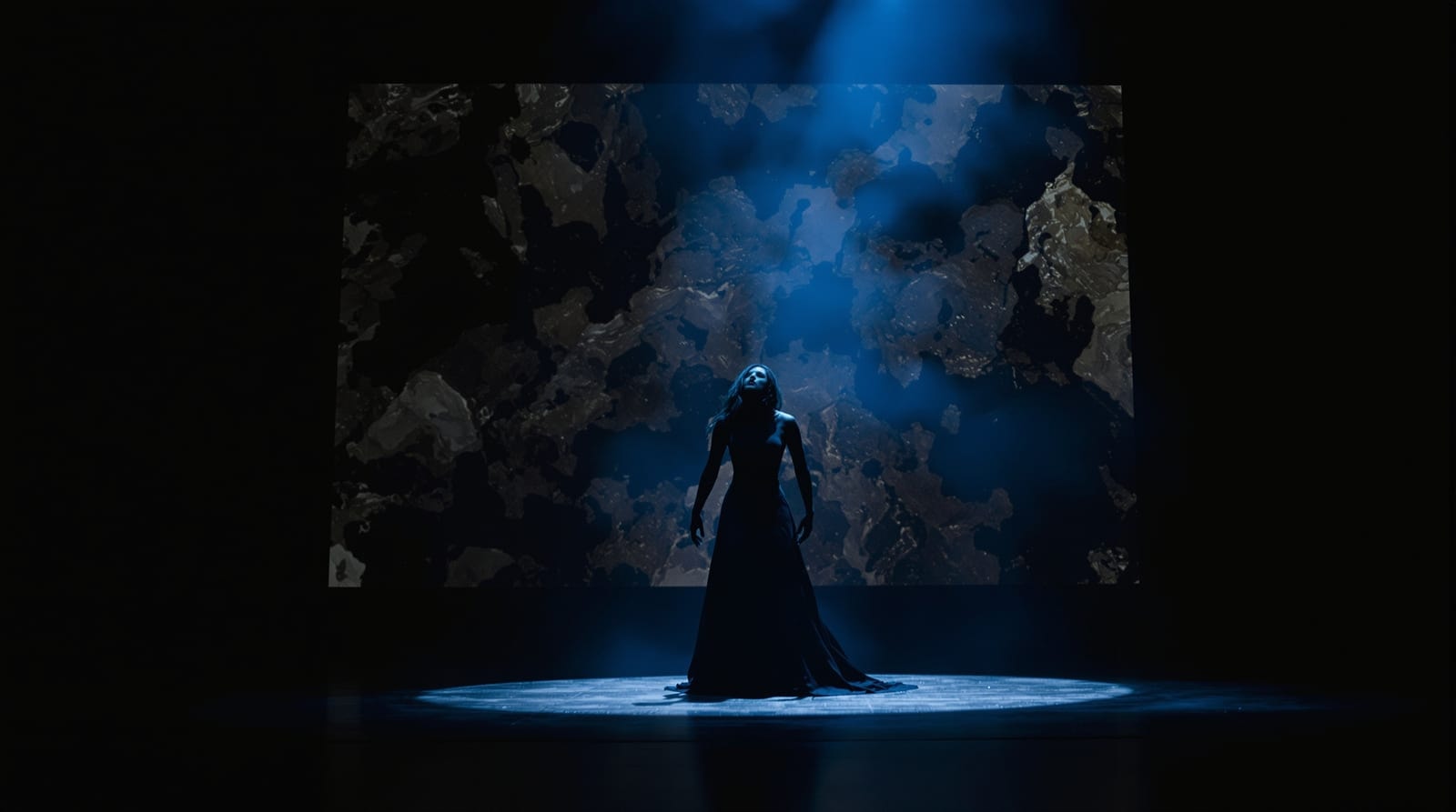A dancer in a long dark costume lit from below in a hazy black box studio with projected texture behind